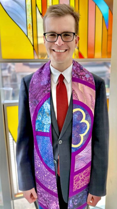 Rev Alex in front of the stained glass panel in the Boardroom